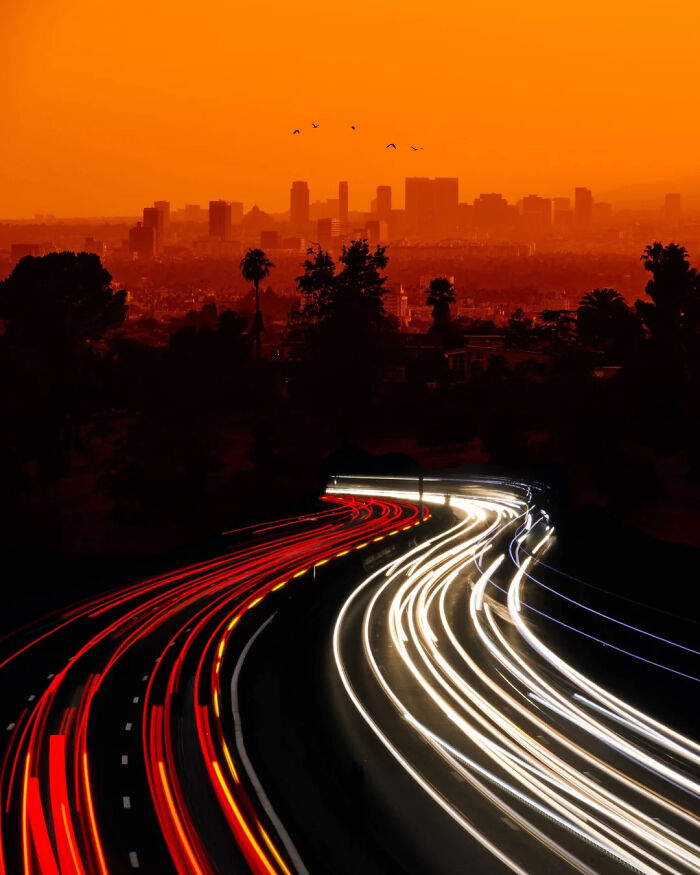 Orange sky over cityscape with light trails on a highway, showcasing minimalism and mystery by Benjamin Wolf.