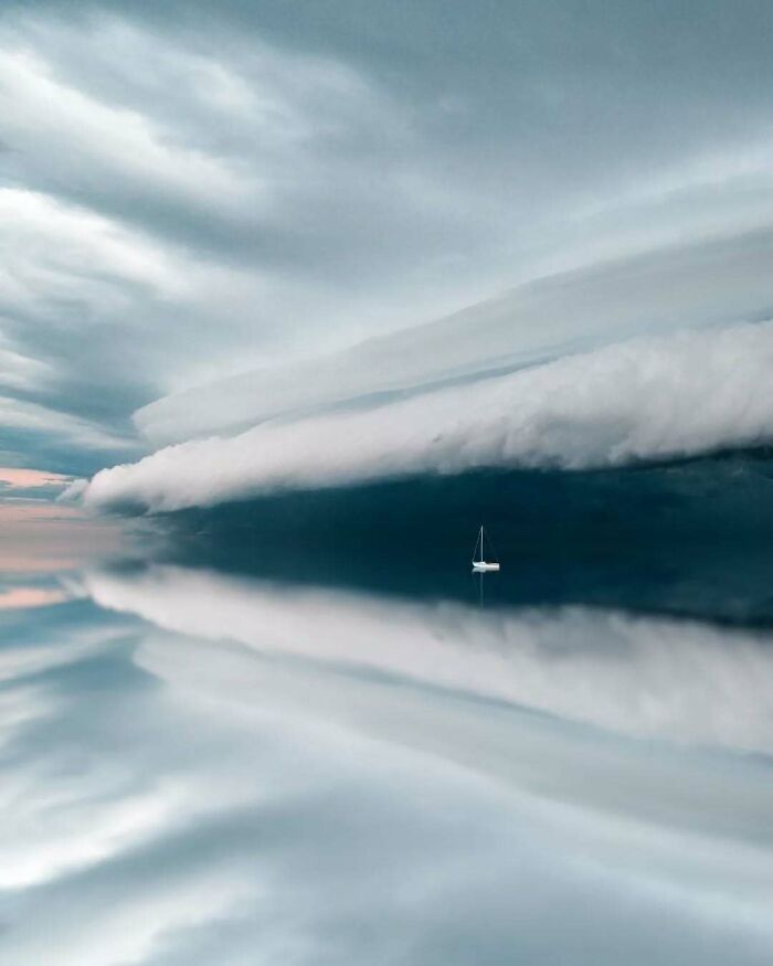 Minimalism and mystery in surreal photo of a sailboat on calm waters under dramatic cloud formations.