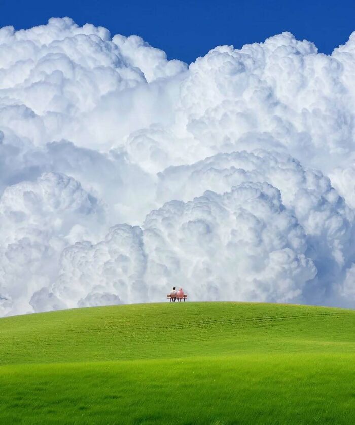 A small figure sits on a bench in a vast green field under towering, surreal clouds, embodying minimalism and mystery.