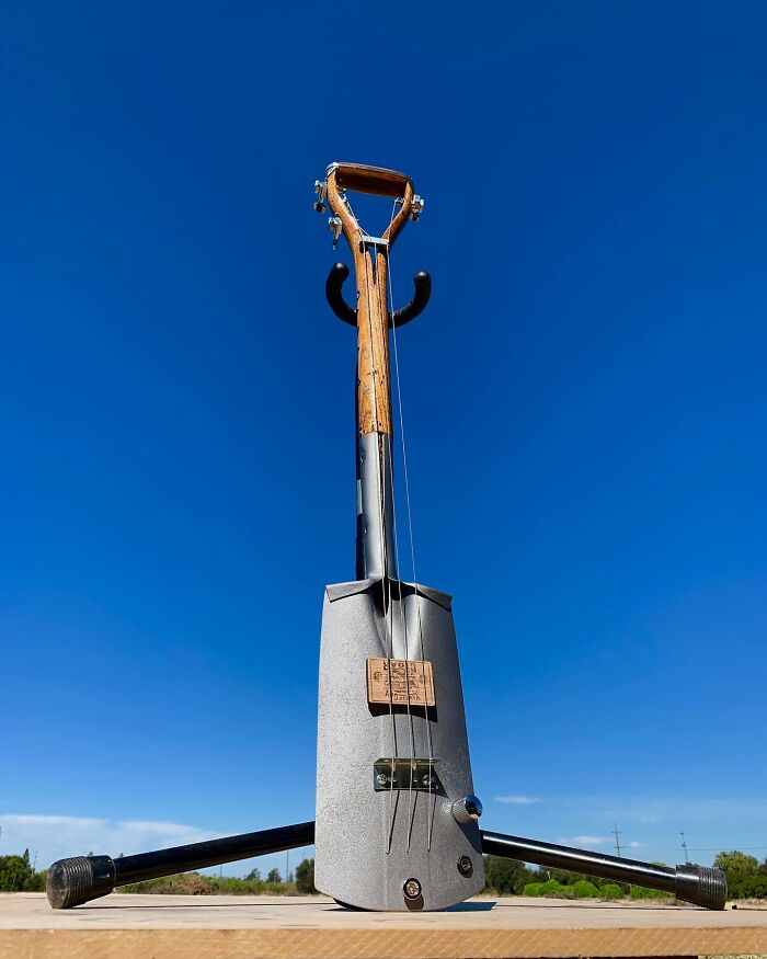 Guitar crafted from unexpected materials, featuring a shovel handle and metal body, set against a clear blue sky.