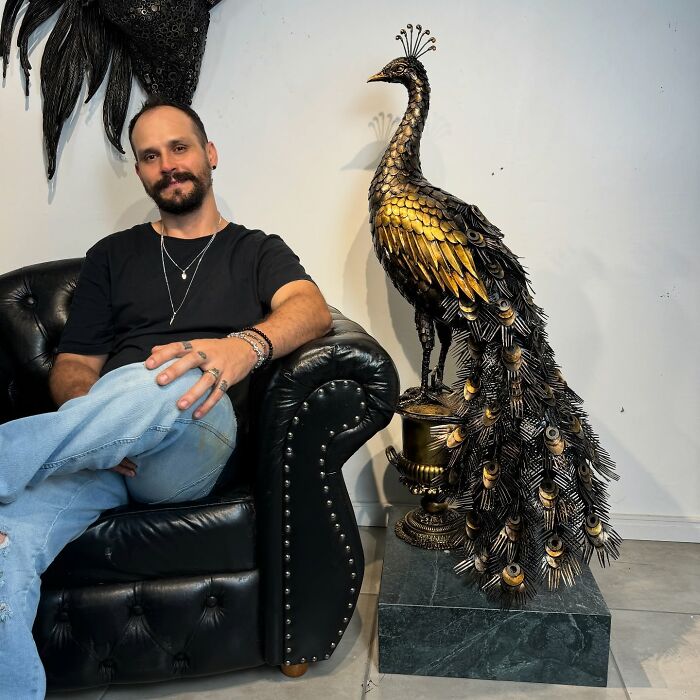 Artist sitting next to a large peacock sculpture made from scrap metal, showcasing intricate and incredible craftsmanship.
