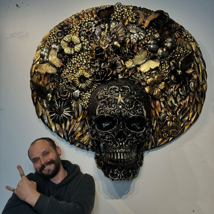 Artist posing with intricate scrap metal skull sculpture adorned with flowers and butterflies.