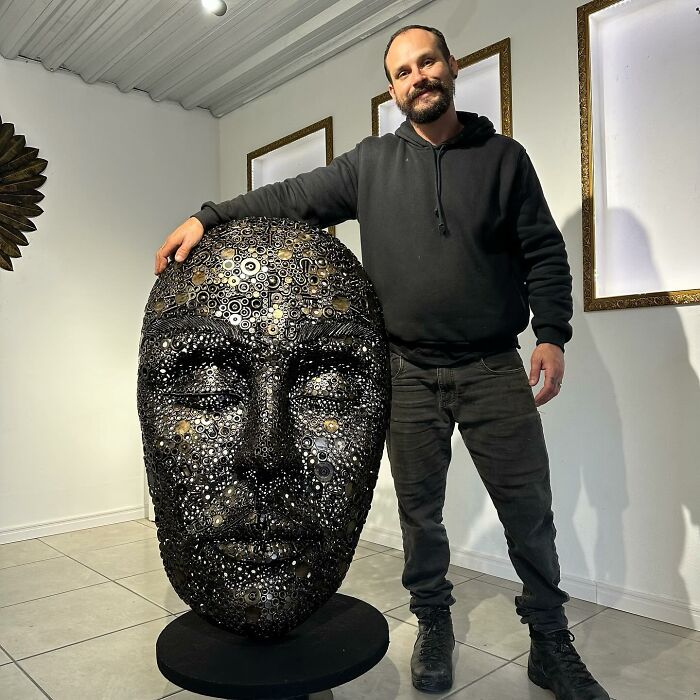 Artist standing next to a large scrap metal sculpture of a face in a gallery setting.