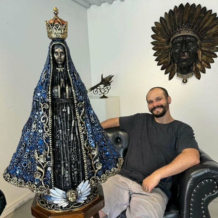 Artist with a scrap metal sculpture of a robed figure, seated in a room with decorative art on the wall.