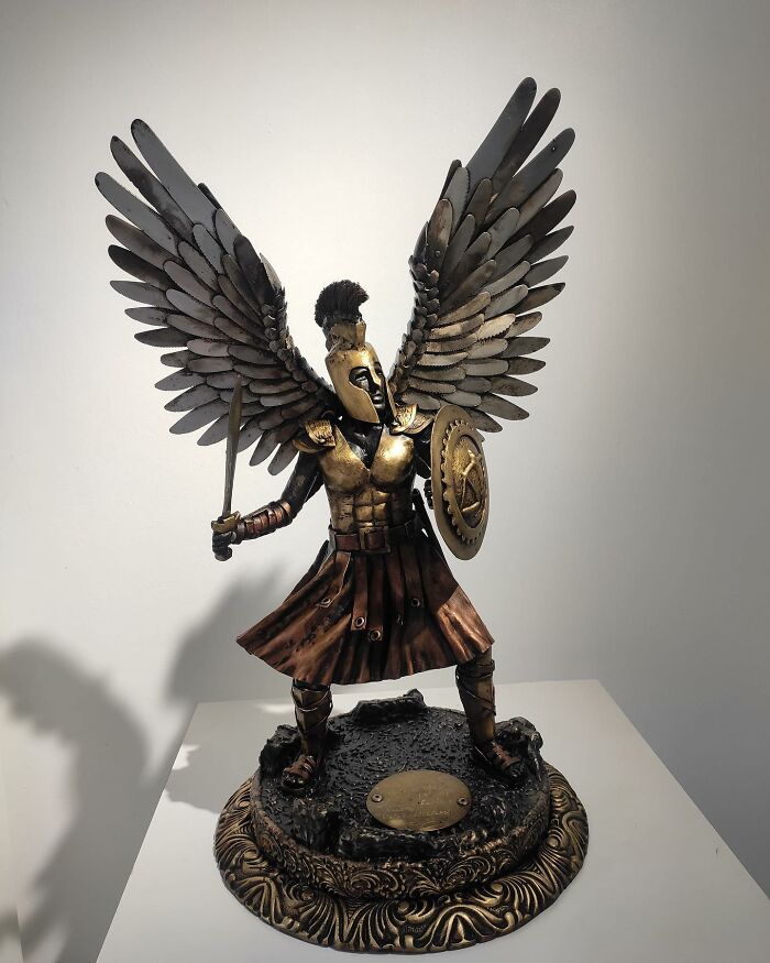 Scrap metal sculpture of a warrior with wings, holding a sword and shield, standing on a decorative base.