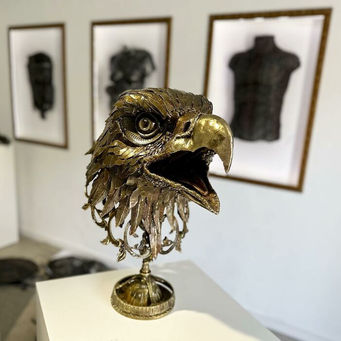 Scrap metal sculpture of an eagle head, displayed in a gallery with framed artworks in the background.