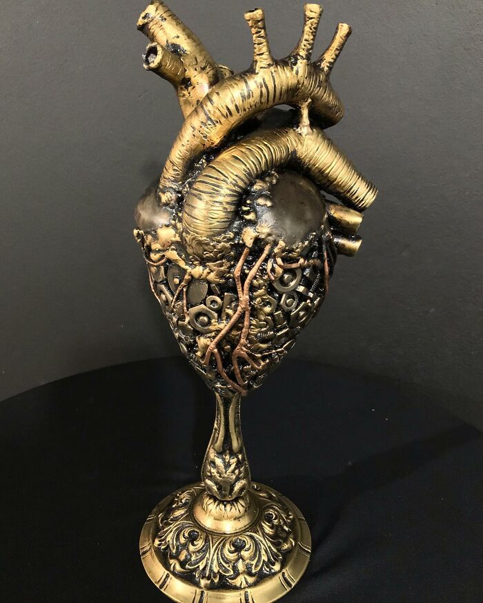 Scrap metal sculpture of a human heart, intricately designed with gold and bronze accents, displayed on a black background.