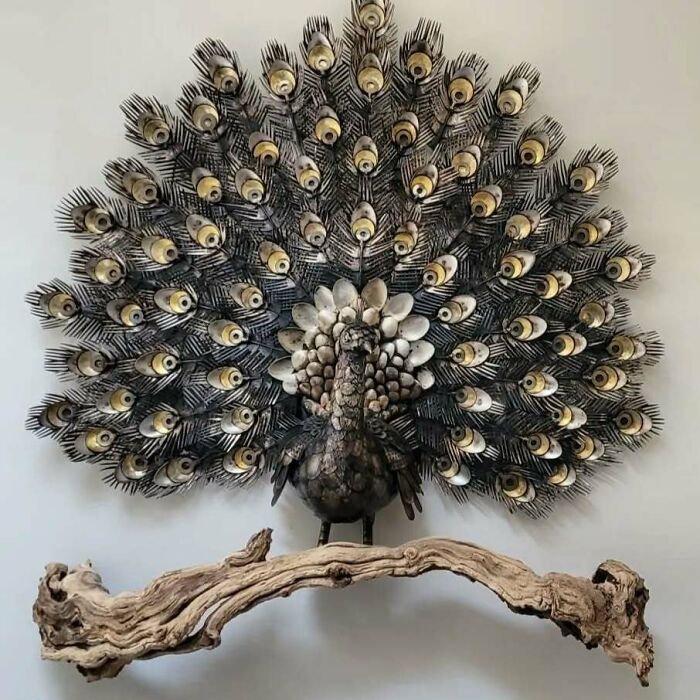 Scrap metal sculpture of a peacock with detailed feathers displayed above a wooden branch.