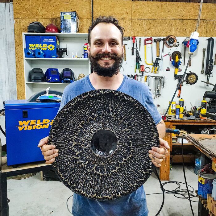 Artist in workshop holding a large circular scrap metal sculpture with tools and equipment visible in the background.