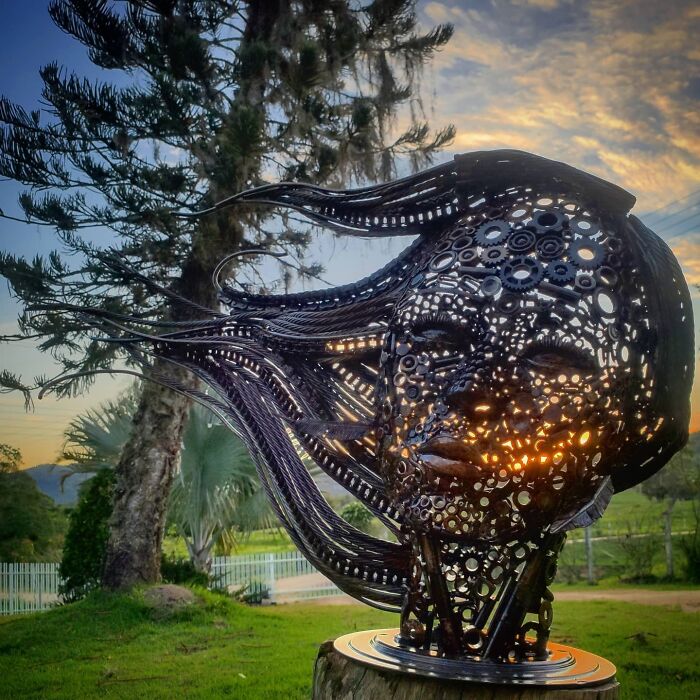 Scrap metal sculpture of a face with flowing hair, set against a sunset and tree background.