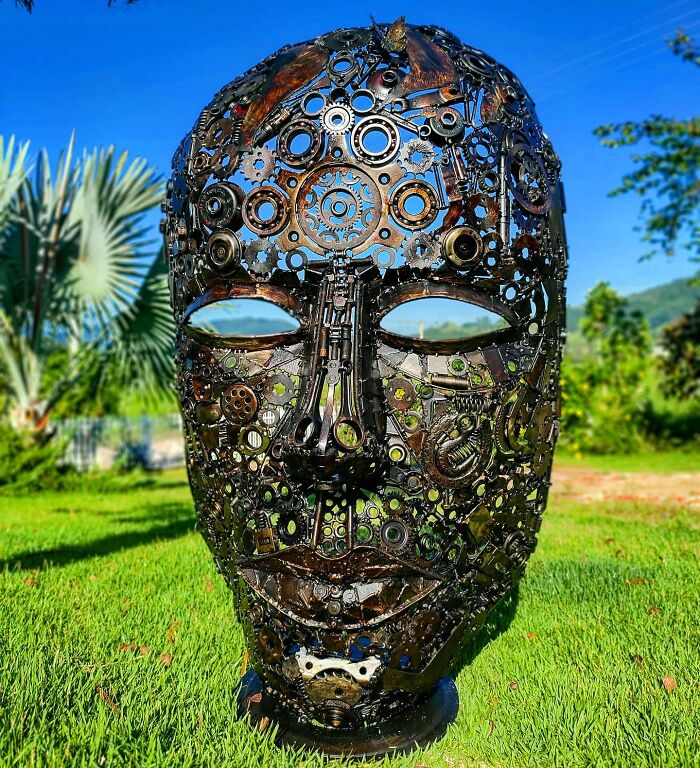 Scrap metal sculpture of a human face, detailed with gears and parts, displayed outdoors on grass.