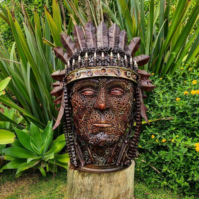 Scrap metal sculpture resembling a face with intricate details, set against a lush garden backdrop.