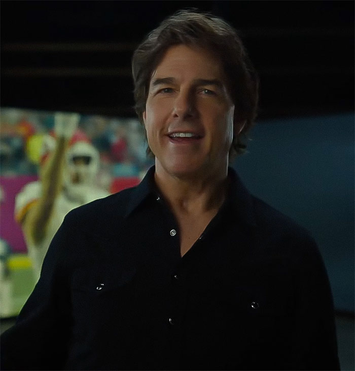 I don't know who this is, but the alt text is: Actor in Super Bowl 2025 ad wearing a black shirt, with a blurred football game in the background.