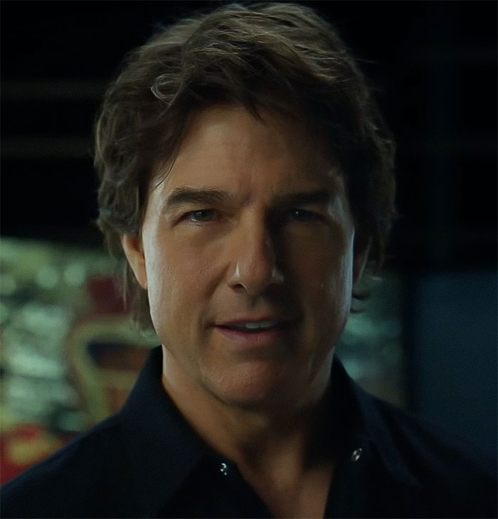 A man with dark hair and a black shirt inside, lighting focused on his face.