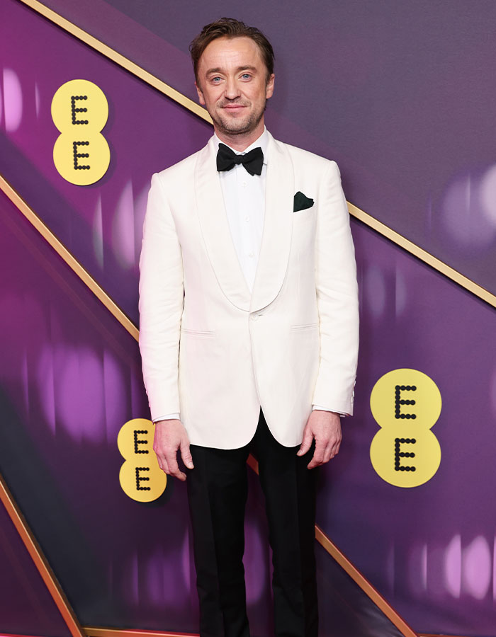 Individual in a white tuxedo with black bow tie at Bafta red carpet, posing against a purple backdrop with yellow logos.