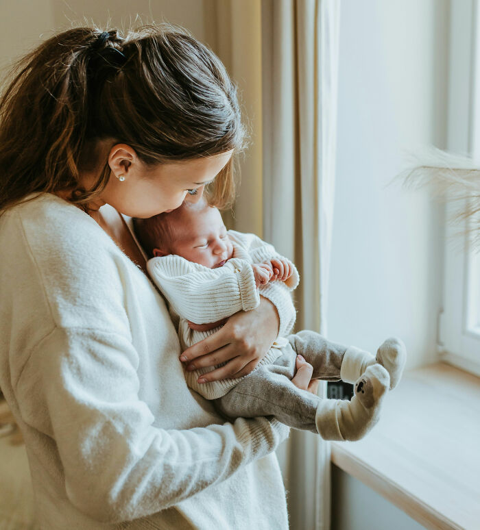 Woman holding a baby by a window, wearing a cozy sweater, showcasing a tender moment.
