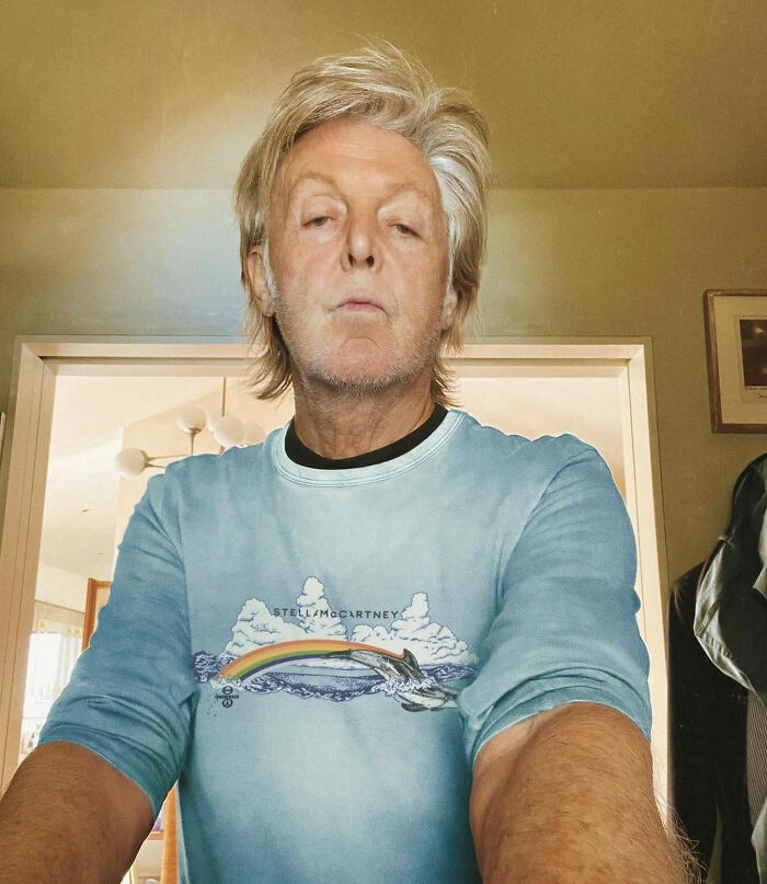 Celebrities: Person wearing a blue T-shirt with a rainbow design, standing indoors.