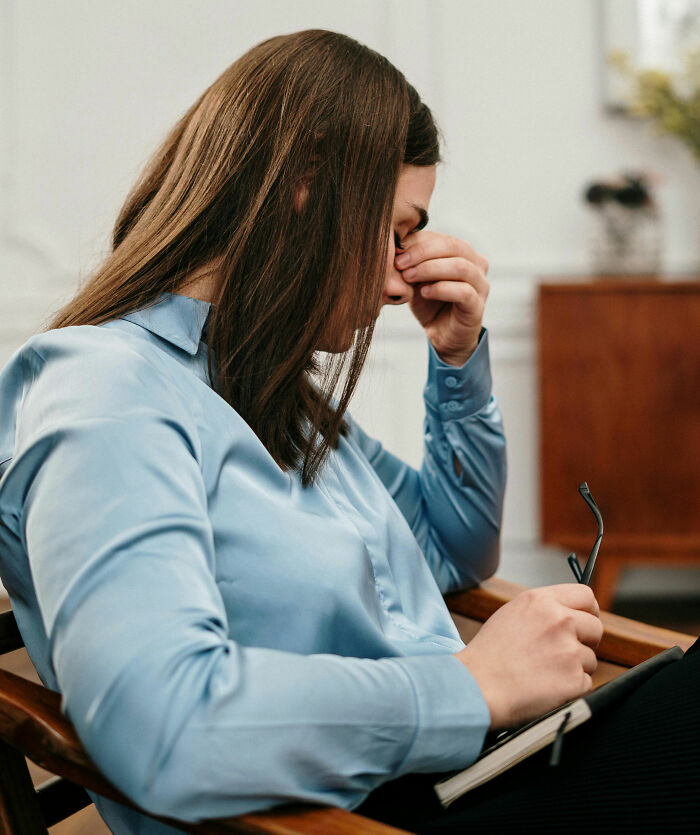 Woman in a blue shirt sitting with a thoughtful expression, holding glasses and a notebook, contemplating offensive comments.