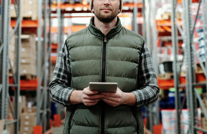 Man in a warehouse using a tablet, wearing a green vest and plaid shirt, surrounded by shelves and boxes.