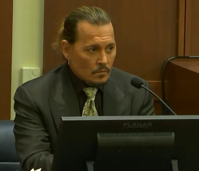 A celebrity with a tied-back hairstyle testifying in a courtroom, wearing a black suit and paisley tie.