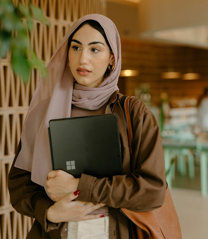 Young woman in a hijab holding a laptop, conveying thoughtful expression.