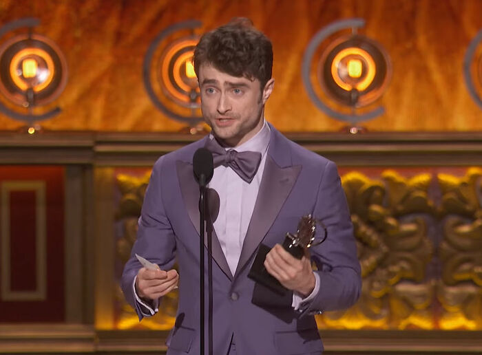 Celebrity holding an award in a purple suit on stage, speaking into a microphone.