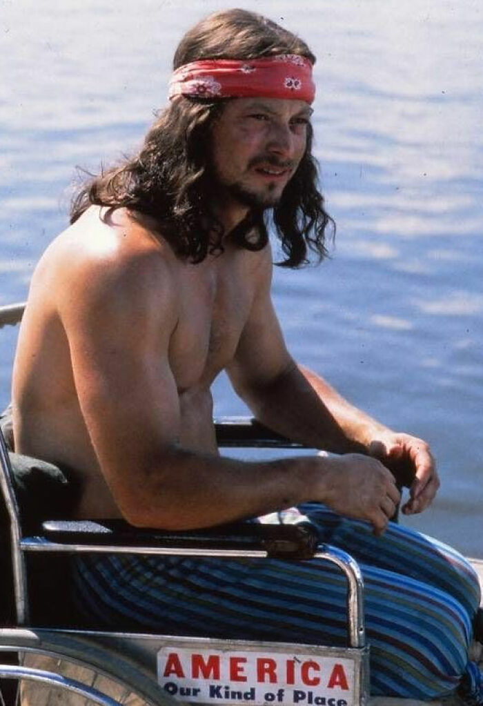 Shirtless man with long hair and headband sitting in a wheelchair near water, with "America Our Kind of Place" sign visible.