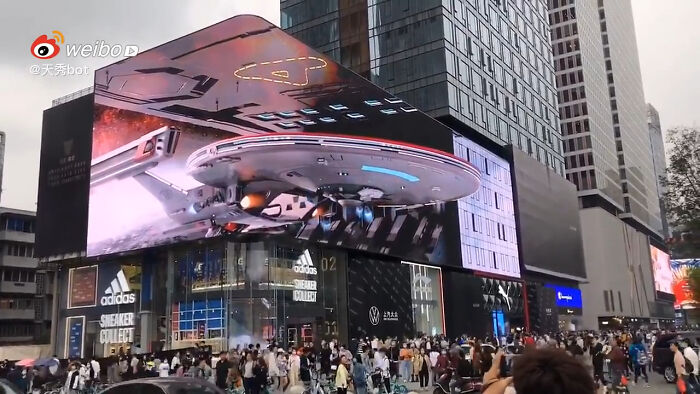 3D spaceship on large screen captivates tourists in bustling city, showcasing an interesting experience at a tech intersection.
