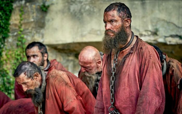 Bearded man in chains, wearing worn red clothing, surrounded by others, illustrating rules from one individual's actions.