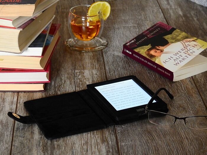 E-reader and glasses on wooden table with stacked books and a tea cup.