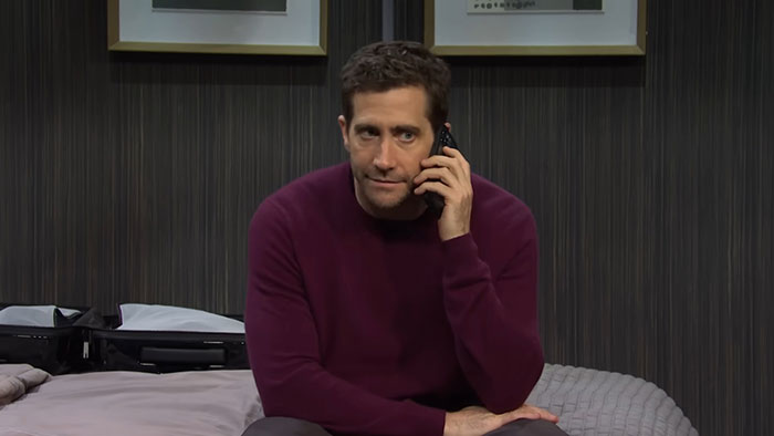 Actor in a maroon sweater sitting on a bed, talking on the phone.