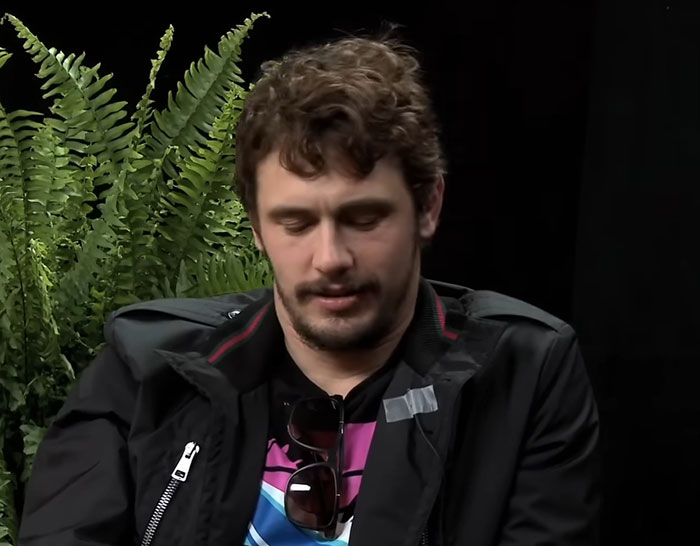 Man with curly hair and beard, sitting in front of ferns, wearing a black jacket and sunglasses hanging on his shirt.