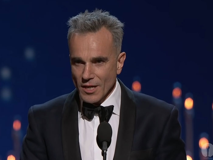 Actor in a tuxedo speaking at an event, with candles in the background.