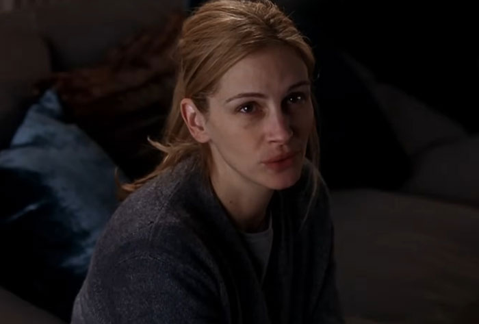 An actor sitting on a couch, wearing a gray sweater, with a neutral expression.
