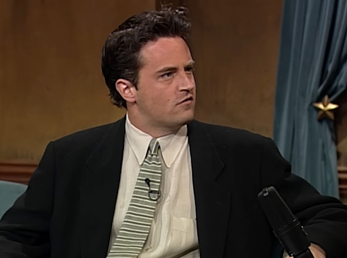 Actor in a suit and tie on a talk show set, looking to the side.
