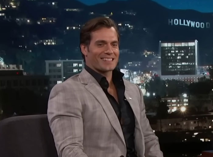 A smiling actor in a grey suit during a TV interview, with the Hollywood sign in the background.
