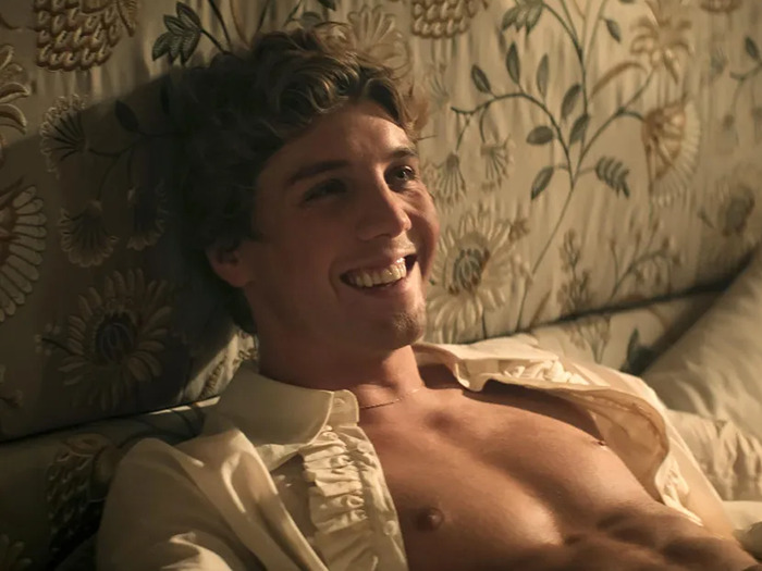 Lukas Gage smiling, reclining shirtless on a patterned couch, demonstrating natural approach without prosthetics.