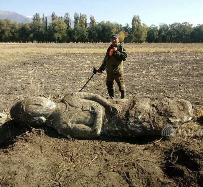 A person stands next to an ancient artifact sculpture in an open field, holding a shovel.
