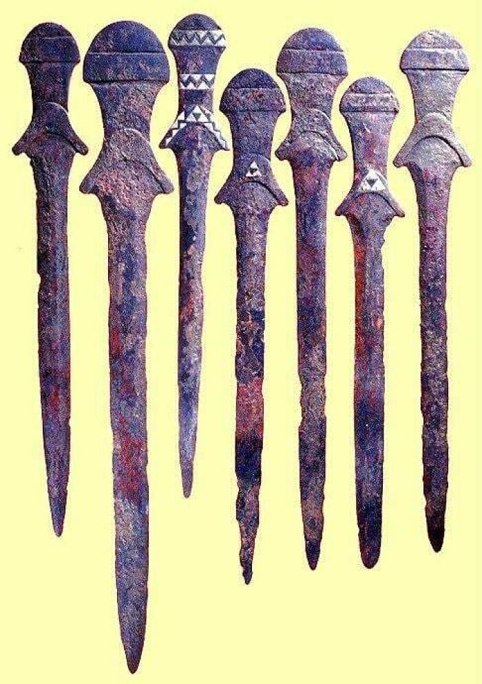 Ancient artifacts: seven rusted daggers from early civilizations displayed vertically against a beige background.