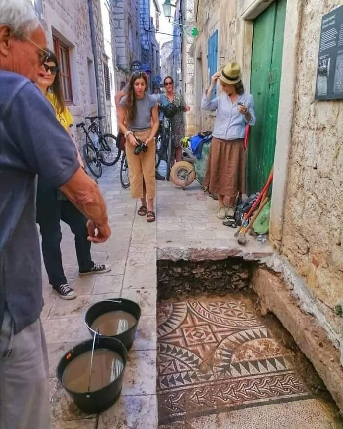 People observing ancient mosaic artifacts in a narrow street excavation site.