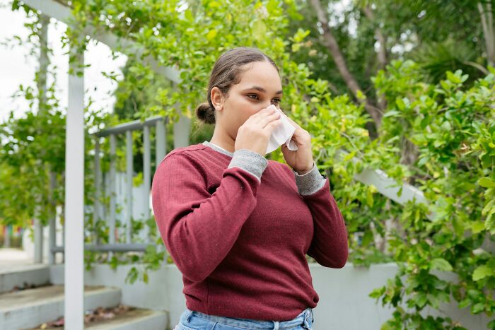 Person experiencing culture shock, wearing a red sweater, using a tissue in a green, outdoor setting.