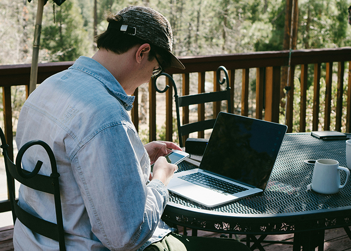 Person using a phone and laptop on a cabin deck with trees in the background, highlighting family cabin use. Person using a phone and laptop on a cabin deck with trees in the background, highlighting family cabin use.