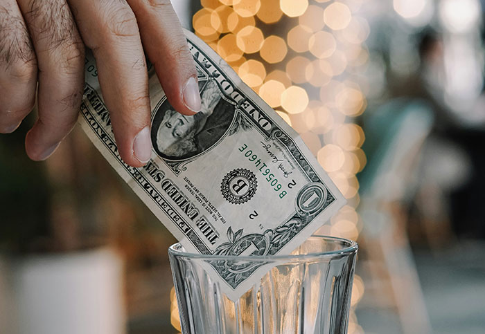 Hand placing a dollar bill in a tip jar, sparking a debate on tipping culture.