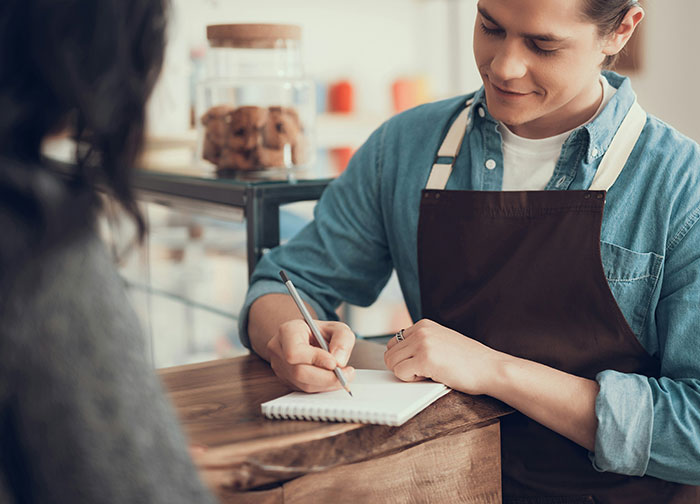Server in a blue shirt and apron writing in a notepad, sparking debate on customer tipping practices.