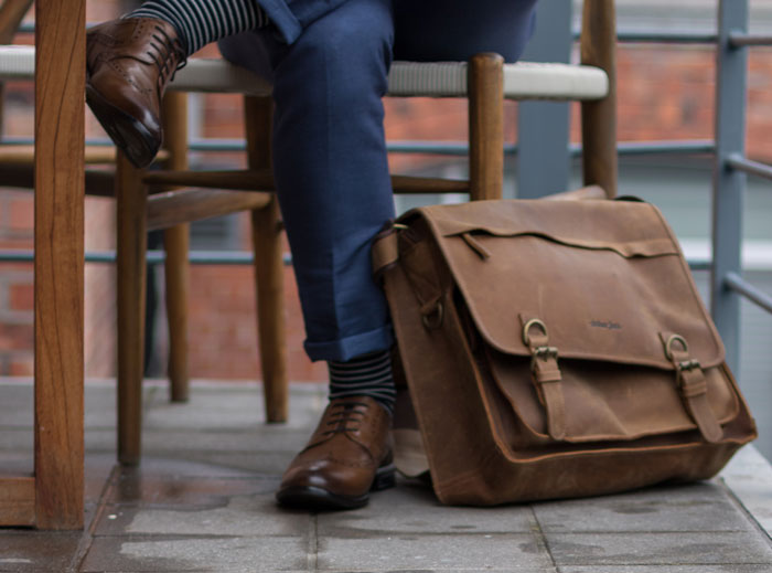 Man sitting on a balcony with brown leather bag, wearing blue pants and brown shoes.