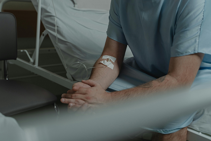 Man in hospital attire sitting beside a bed, with an IV bandage on his arm.