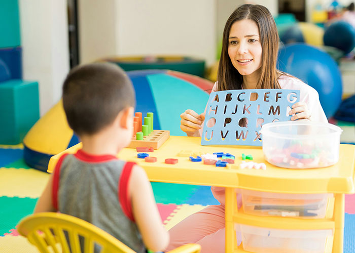 Teacher showing alphabet letters to a young boy in a colorful classroom setting.