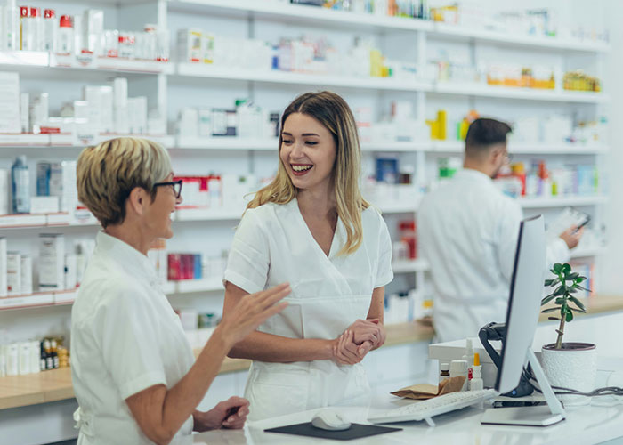 Pharmacy staff smiling and talking near a computer, surrounded by shelves of medicine.