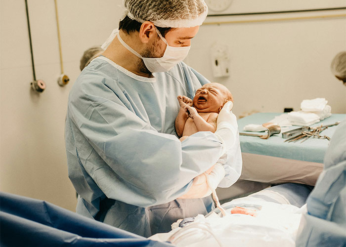 A doctor in scrubs and mask holds a newborn baby in a hospital delivery room.