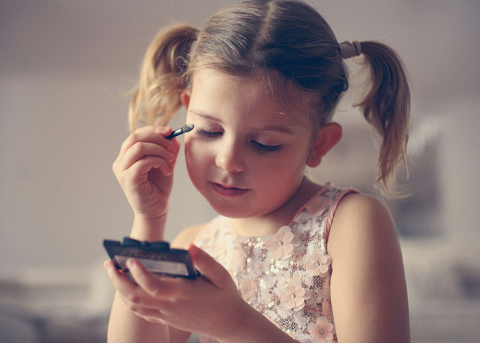 Young girl with pigtails applying makeup, wearing a floral dress, embodying innocence and creativity.